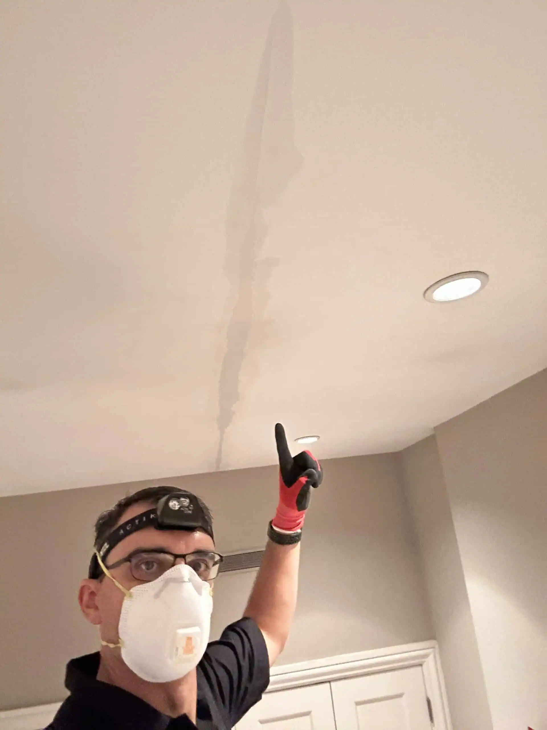 A person wearing a mask, headlamp, and gloves points to a long crack in a white ceiling, indicating possible water damage. They stand in a softly lit room with neutral-colored walls, perhaps considering hiring painting services NYC to restore the pristine look.