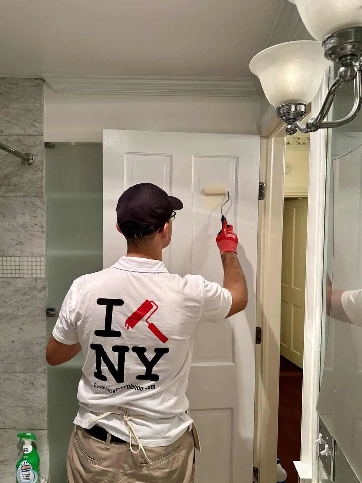 A person wearing a cap and a T-shirt with a paint roller design is painting a white door in a room, exemplifying the skill of top painting contractors NYC. Cleaning supplies are visible on the left. The room has tiled walls and a light fixture.