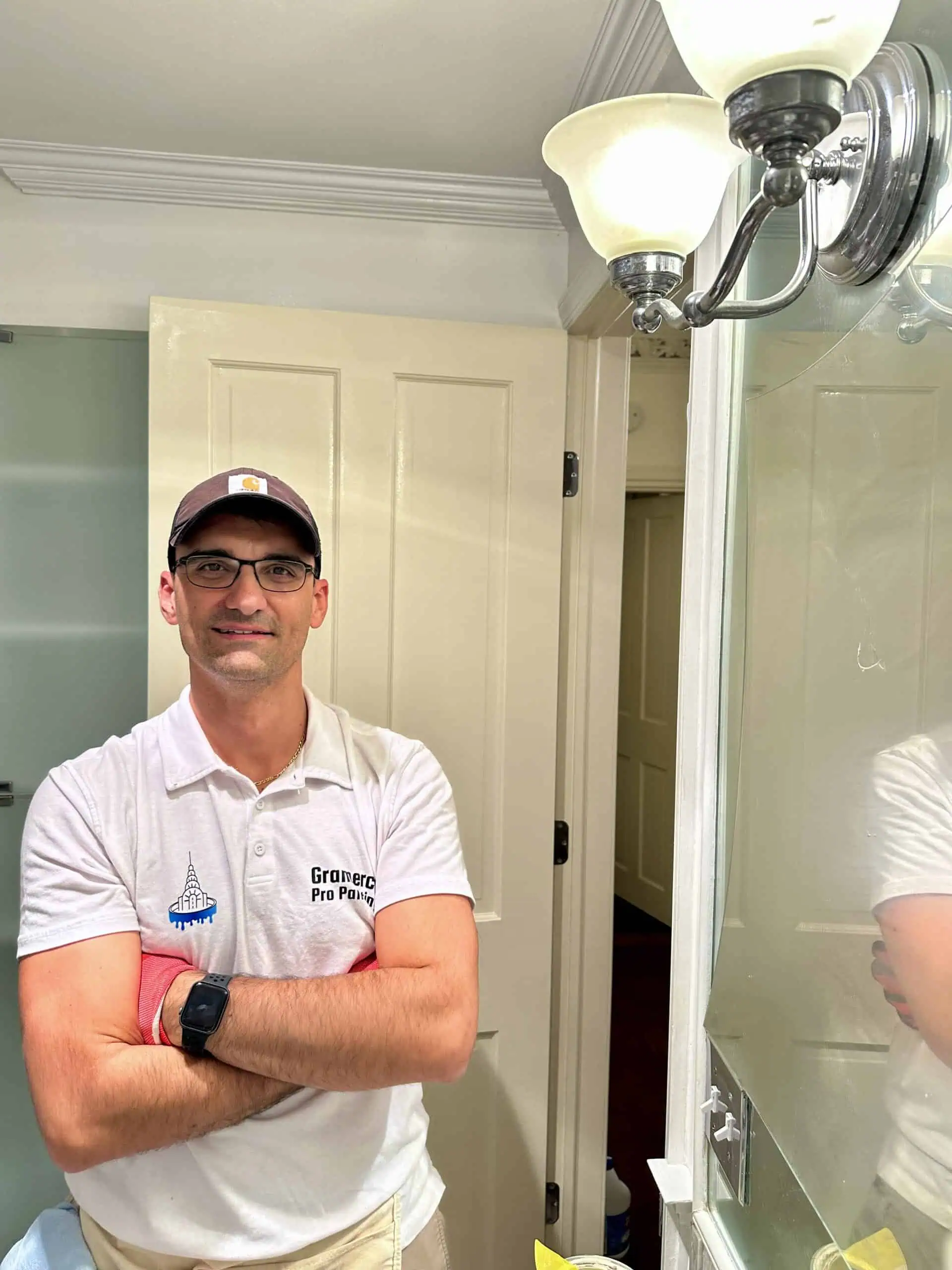 A person wearing a cap and glasses stands with crossed arms in a bathroom, where the pristine walls clearly reflect the work of top painting contractors NYC. They are in a white polo shirt with a logo and text, wearing a black watch beneath the wall-mounted light fixture.