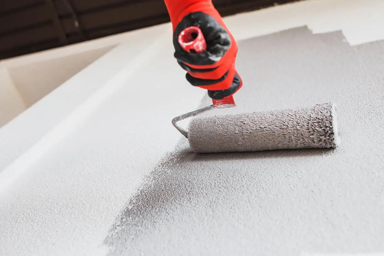 A person in red gloves uses a paint roller to apply light gray paint on a white wall. The roller is partially covered, leaving a mix of freshly painted and unpainted sections—a scene typical for skilled painting services NYC trusts for impeccable results.