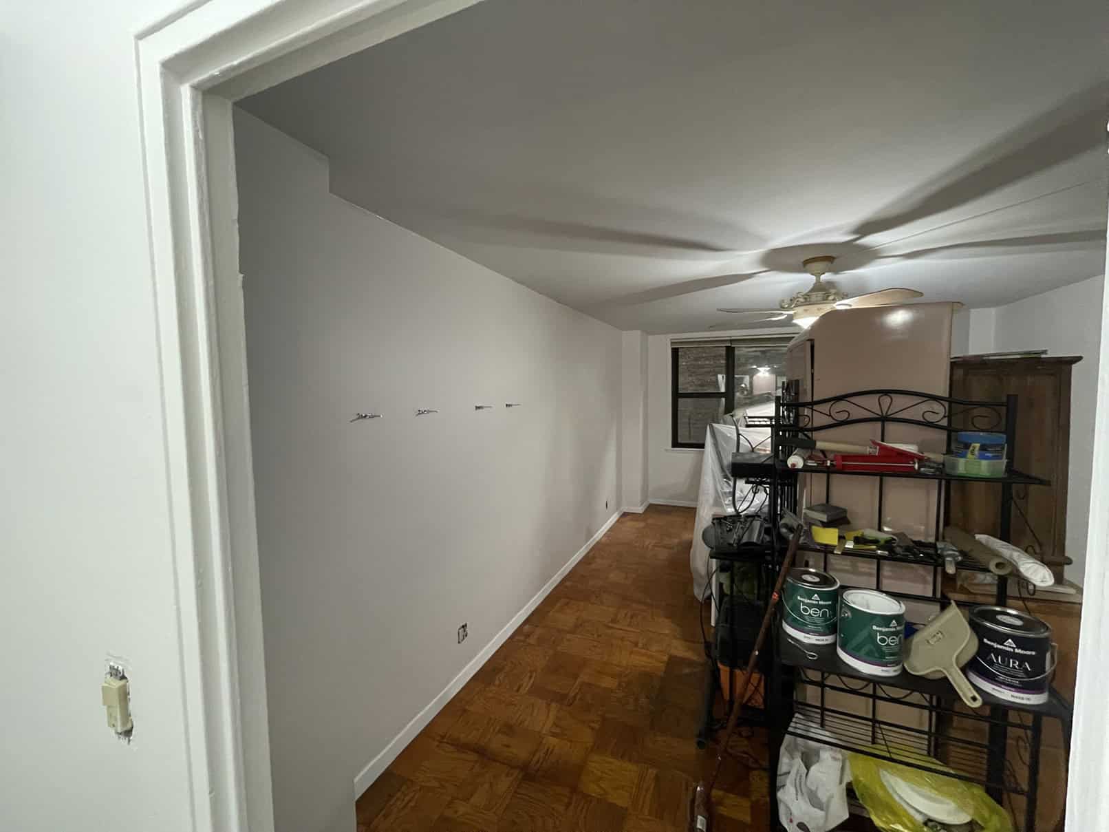 A narrow room with a white wall on the left, wooden floor, and shelves holding paint cans from top painting contractors NYC. A ceiling fan spins quietly above, while a window offers an outside view at the far end. Hooks are installed on the walls, showcasing efficient space usage for painting services NYC.