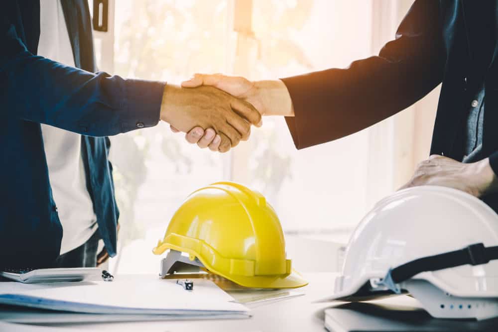 Two people shaking hands over a table with yellow and white construction helmets and documents, symbolizing agreement or partnership. Sunlight filters through a window in the background.