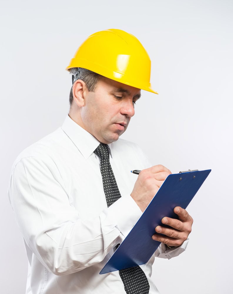 A man wearing a yellow hard hat and a white dress shirt writes on a blue clipboard. He is holding a pen and appears focused on his task. The background is a plain white.